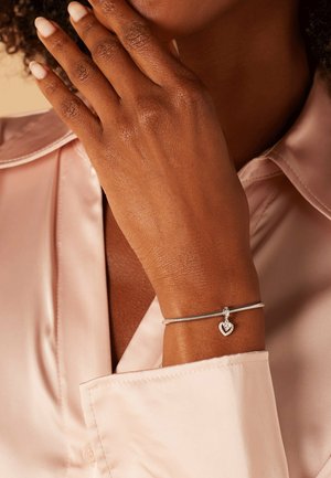 Silbernes Armband mit einem Herzen-Anhänger, verziert mit kleinen klaren Steinen, getragen an einer Hand, die neben einem weichen rosa Satinhemd ruht.