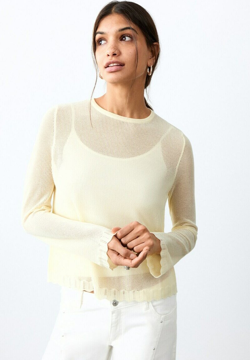 Femme aux cheveux foncés attachés en queue de cheval basse, portant un haut à manches longues en maille crème et un pantalon blanc, regardant légèrement sur le côté, les mains jointes.