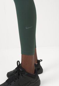 Leggings atléticos verdes con un diseño ajustado y un pequeño logo gris de Nike, combinados con zapatillas negras con cordones sobre un fondo claro.