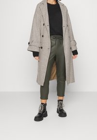 Manteau à double boutonnage à carreaux beige et noir, associé à un pull en maille noir et un pantalon tapered vert olive. Complété par des bottes à lacets noires.