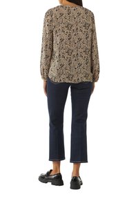 Langarmige, gebloemde blouse in beige en zwart, gecombineerd met donkere, cropped jeans. De outfit heeft een ontspannen pasvorm en glanzende zwarte schoenen.