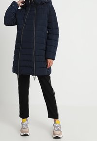 Marinblå puffjacka med quiltad design, framsidan med dragkedja och snörning i huvan, i kombination med svarta träningsbyxor och sneakers med gula detaljer.