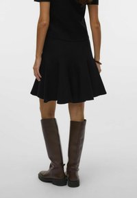 Jupe noire évasée en tissu doux, associée à de longues bottes en cuir marron. La jupe présente une surface légèrement texturée avec des détails de plis.