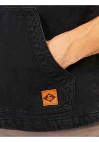 Czarna dżinsowa kurtka z boczną kieszenią, z beżową metką znaku firmowego i teksturowanym szwem. Materiał jest wytrzymały i ma gładkie wykończenie.