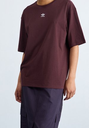 Persoon draagt een donker kastanjebruine Adidas T-shirt met logo op de borst en donkere gestreepte cargobroek, staande tegen een effen achtergrond.