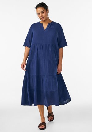 Moteris dėvi laisvą, sluoksniuotą tamsiai mėlyną midi suknelę trumpomis rankovėmis ir V formos iškirpte, žingsniuoja pirmyn vilkėdama rudas plokščias sandalus.