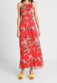 Maxikjole i rødt stof med et blomsterprint, der har store blade og tropiske blomster. Halterneck-design, flydende facon, fremhævet med et bælte.