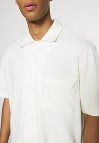 Biała polo koszulka z kołnierzykiem, wykonana z miękkiego materiału, z kieszonką na piersi i zapięciem na guziki. Gładka tekstura i klasyczny krój.