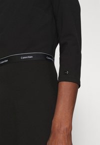 Černé šaty s pasem s logem Calvin Klein. Mají tříčtvrteční rukávy a hladkou, strukturovanou tkaninu. Akcent s logem na manžetě rukávu.