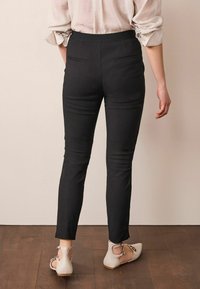 Zwarte slim-fit broeken met een gladde textuur, voorzien van twee achterzakken en een hoge tailleband, gecombineerd met lichtgekleurde schoenen.