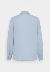 Chemise bleu clair à manches longues avec poignets boutonnés, vue de dos sur fond blanc.