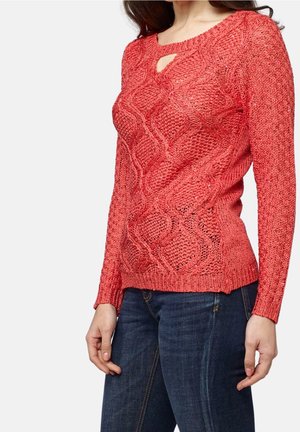 Femme portant un pull en tricot corail texturé avec un petit décolleté en trou de serrure et un jean bleu foncé sur un fond uni.