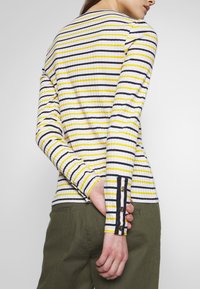 Långärmad ribbad topp i vitt med gula och svarta horisontella ränder. Har knappe detaljer vid ärmsluten och en figurnära silhuett.