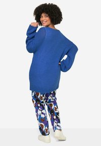 Blauwe oversized gebreide sweater met wijde mouwen, gecombineerd met gestreepte broeken met gezichtontwerpen in paars en bruin op een witte achtergrond.