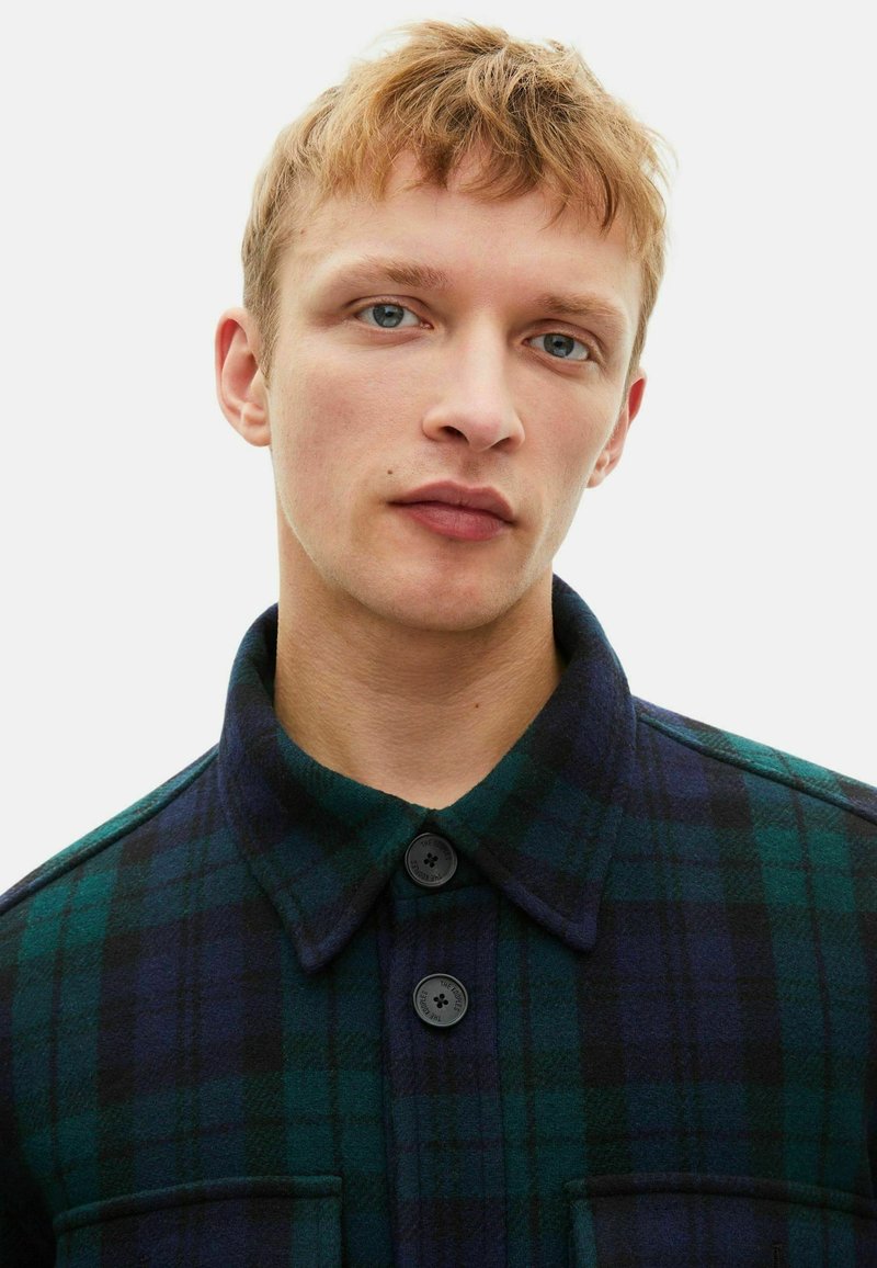 Junger Mann mit kurzen blonden Haaren, der eine dunkelgrüne und marineblaue karierte Jacke mit Knöpfen vor einem schlichten weißen Hintergrund trägt.