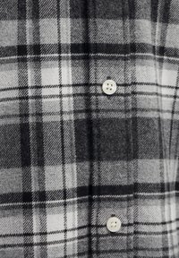 Rutete flanellskjorte i svart, grått og hvitt. Har store rutemønstre, teksturert stoff og en hvit knapp ved kragen.
