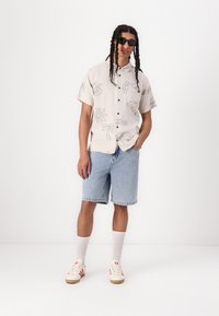Camicia a maniche corte color crema con motivi neri, pantaloni shorts in denim blu, calze bianche e sneakers color crema con dettagli rossi. Occhiali da sole neri indossati.