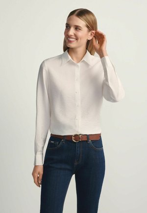 Uśmiechnięta kobieta w białej, długorękawowej koszuli zapinanej na guziki, niebieskich dżinsach i brązowym pasku, dotykająca włosów, stojąca na tle jednolitego, jasnego tła.