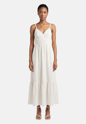 Vrouw staat en draagt een mouwloze witte maxi-jurk met een diepe V-hals en dunne bandjes, gecombineerd met witte sandalen met hak.