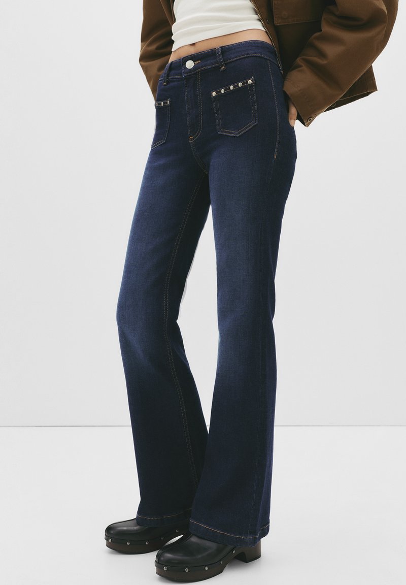 Jeans in denim blu scuro a zampa d'elefante con tasche frontali dotate di borchie argentate e cuciture arancioni a contrasto. Indossati con zoccoli neri spessi.