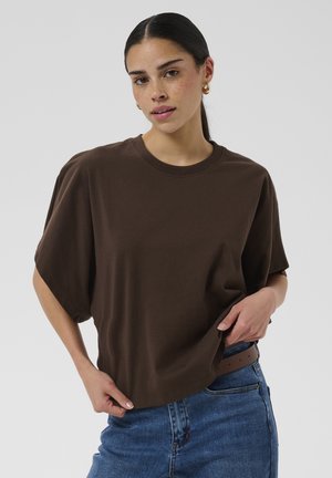 Jeune femme aux cheveux foncés vêtue d’un t-shirt marron oversize et d’un jean bleu, posant avec une main qui ajuste son t-shirt.