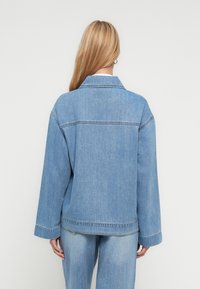 Kvinne med langt blondt hår, iført lys blå denimjakke og jeans, stående med ryggen til betrakteren mot en ensfarget hvit bakgrunn.