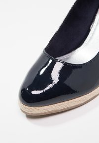 Mitaine compensée en cuir verni bleu marine avec bout pointu, garniture en jute et doublure intérieure lisse. Présente une finition brillante et un design minimaliste.