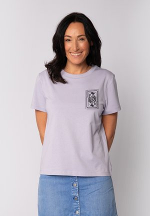 Woman with dark hair smiling, wearing a light purple t-shirt with playing card design on chest and a buttoned blue denim skirt.