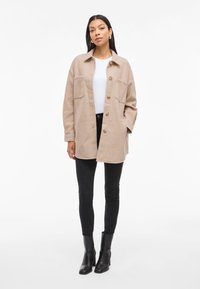 Beige, oversize ingdzseki, galléros nyakkal, két elülső zsebbel és barna gombokkal, fekete skinny farmerral és bokacsizmával párosítva.