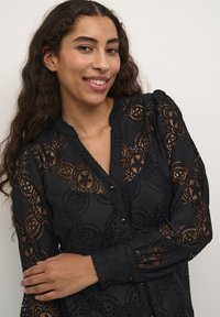 Blusa nera in pizzo con motivi floreali, maniche lunghe e chiusura con bottoni sul davanti. Tessuto testurizzato con dettagli trasparenti e colletto arrotondato.