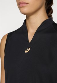 Black sleeveless top featuring a low collar, made of smooth fabric, with a small, gold logo on the chest.