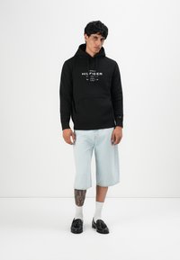 Joven de pie vistiendo una sudadera negra de Tommy Hilfiger, pantalones cortos largos de denim claro, calcetines blancos, mocasines negros con borlas y un tatuaje visible en la pierna.