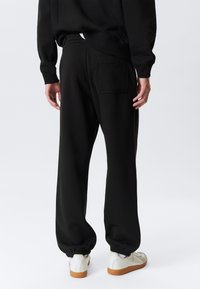 Pantalons de survêtement noirs avec une poche arrière, des poignets élastiques et une coupe décontractée, associés à des baskets blanches. Matériau doux et design minimal.