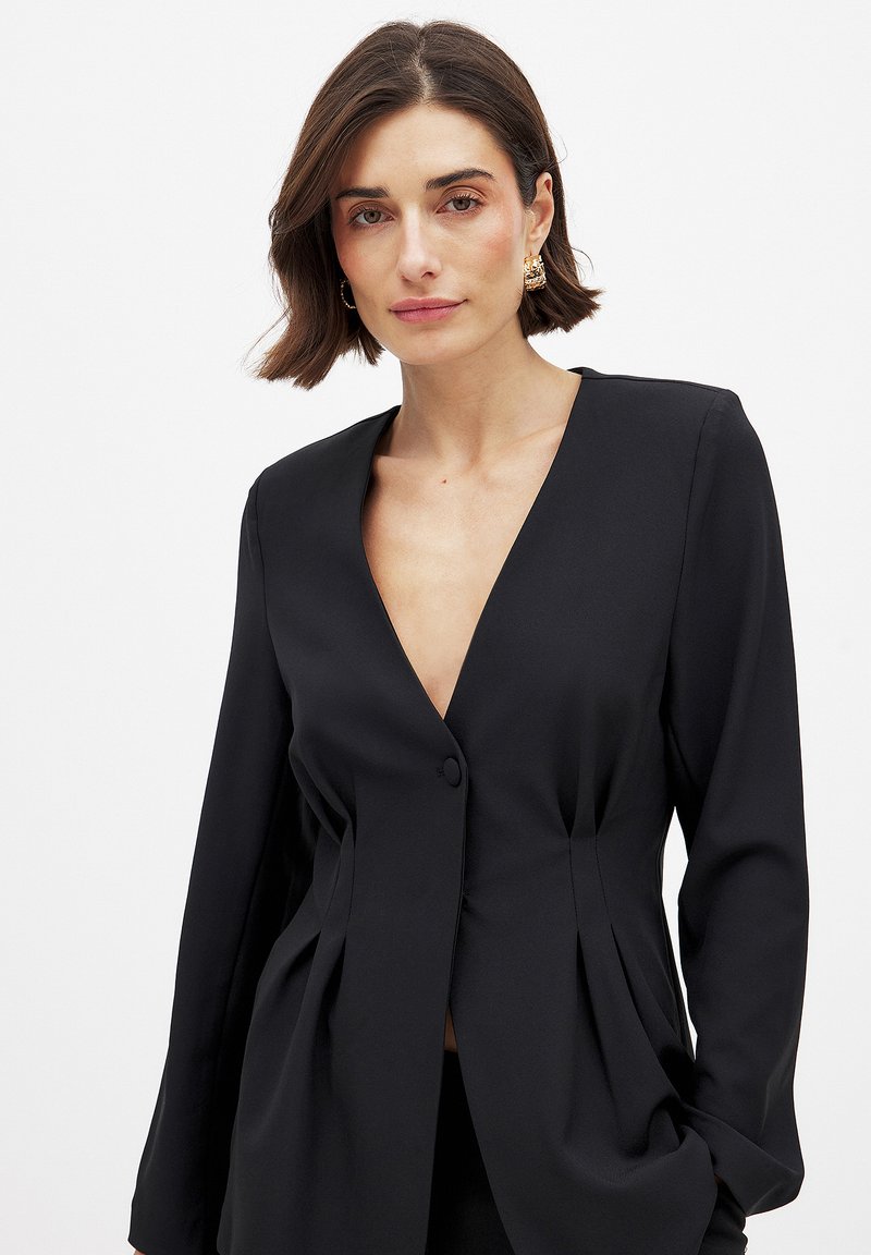 Schwarzer taillierter Blazer mit tiefem V-Ausschnitt, Ein-Knopf-Verschluss und strukturierten Falten an der Taille, gefertigt aus glattem Stoff.