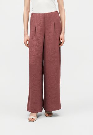 Kobieta ubrana w wysokie, szerokie spodnie o fakturze w kolorze mauve, z plisami z przodu, zestawione z białymi sandałami na obcasie z odkrytymi palcami i białą bluzką.