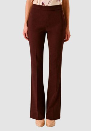 Persona che indossa pantaloni a vita alta, marrone scuro, a gamba dritta e scarpe beige con punta appuntita su uno sfondo grigio uniforme.