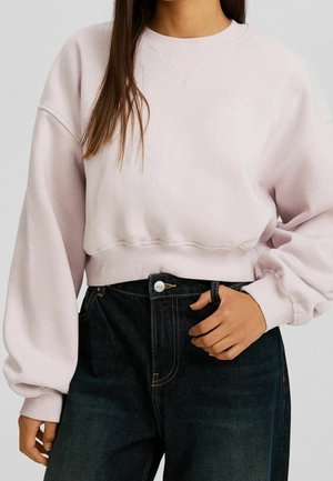 Frau trägt ein hellrosa kurz geschnittenes Sweatshirt mit langen Ärmeln und hoch taillierte dunkelblaue Jeans, Hand ruht nahe dem Bund.