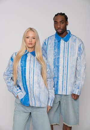 Zwei Models tragen passende blau-weiß gestreifte Hemden mit Knöpfen; eines kombiniert es mit hellen Jeans, das andere mit hellen Denim-Shorts.