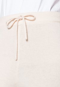 Calças de malha bege clara com um cós canelado, apresentando um cordão de ajuste. Textura suave, vista em close, design minimalista.