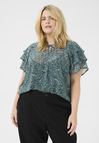 Blusa de manga corta en color verde azulado con mangas con volantes, cuello con lazo y un patrón ondulado en tonos de verde y blanco, combinada con pantalones negros.