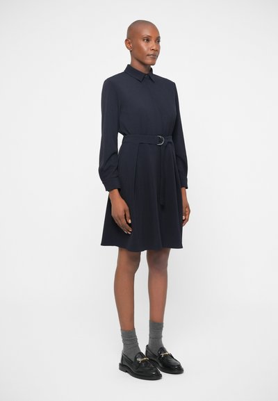 Tamsiai mėlyna suknelė su apykakle, ilgomis rankovėmis ir dirželiu liemens srityje. Pagaminta iš lygaus audinio, su klostėmis sijono krašte.