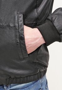 La mano inserita nella tasca di una giacca di pelle nera perforata con polsini a costine, abbinata a jeans in denim blu.