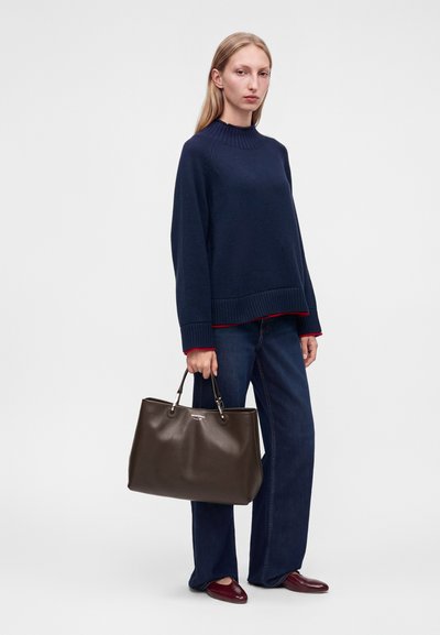 Femme portant un pull bleu marine et un jean, tenant un grand sac à main marron foncé, debout devant un fond uni.