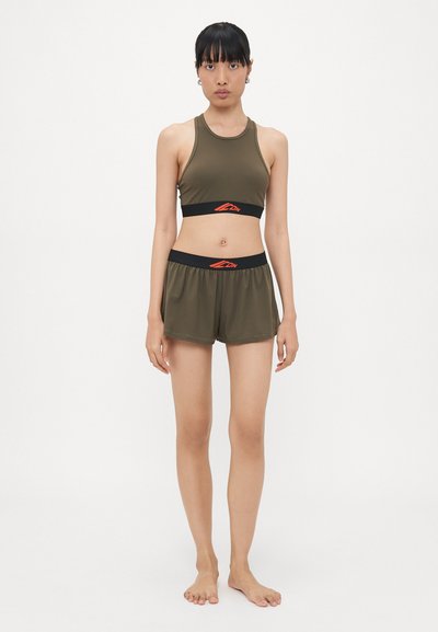 Woman standing barefoot wearing olive green sports bra and matching shorts with black waistband and red logo on plain white background.