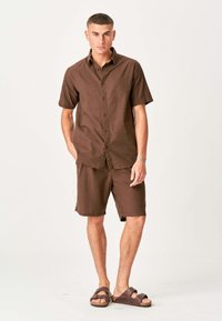 Ung mand står med hænderne i lommerne iført en brun kortærmet skjorte med knapper, matchende shorts og brune sandaler mod en neutral baggrund.