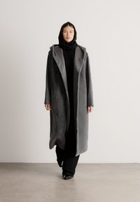 Dlhý sivý kabát s textúrou a kapucňou, s gombíkovou prednou stranou a širokými rukávmi, nosený cez čierny outfit. Hladký povrch s jemným pocitom.