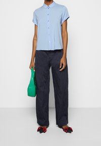 Chemise bleu clair à manches courtes avec boutons, pantalons larges foncés, sac fourre-tout vert et chaussures plates noires ornées de motifs floraux rouges.