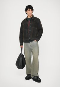 Giacca di jeans nera sopra una maglietta grafica grigia, pantaloni baggy verdi chiari, scarpe slip-on nere con ricamo, mentre tiene una borsa nera.