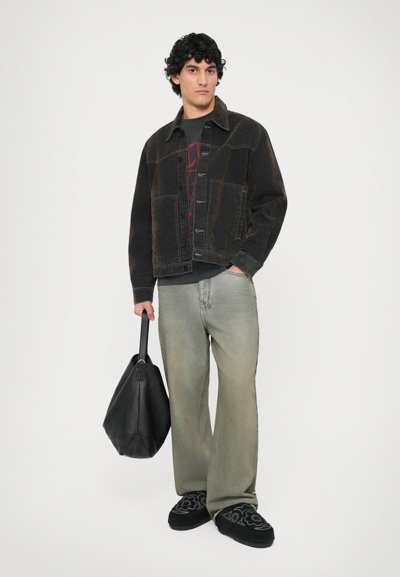 Chaqueta de mezclilla negra sobre una camiseta gráfica gris, jeans holgados verde claro, zapatos negros de lona bordados, sosteniendo un bolso negro.