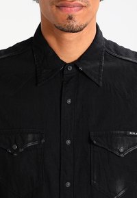 Camicia in denim nero con colletto, due tasche frontali con chiusura a bottone e delicati dettagli scuciti sui bordi.